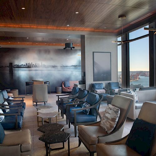 The image shows a modern lounge with multiple seating arrangements, large windows offering a scenic view of water and a Ferris wheel in the distance.
