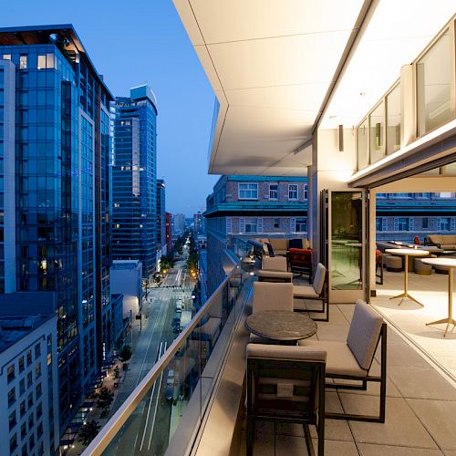 A modern city view during dusk from a balcony with glass railings, outdoor seating, and tables, adjacent to a well-lit indoor area.