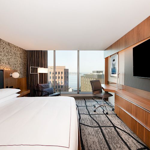 A modern hotel room with a bed, wall-mounted TV, desk, chair, and floor-to-ceiling windows offering a city and water view in the background.