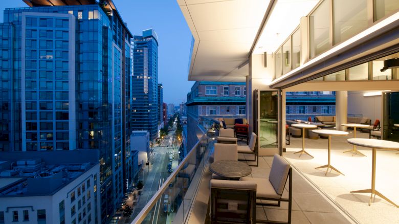 A rooftop terrace overlooking a city with modern high-rise buildings, tables, and chairs, under evening city lights.