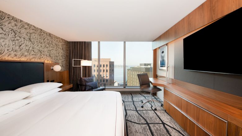 A modern hotel room with a large bed, a desk, a chair, a wall-mounted TV, and a window with a cityscape view in the background.