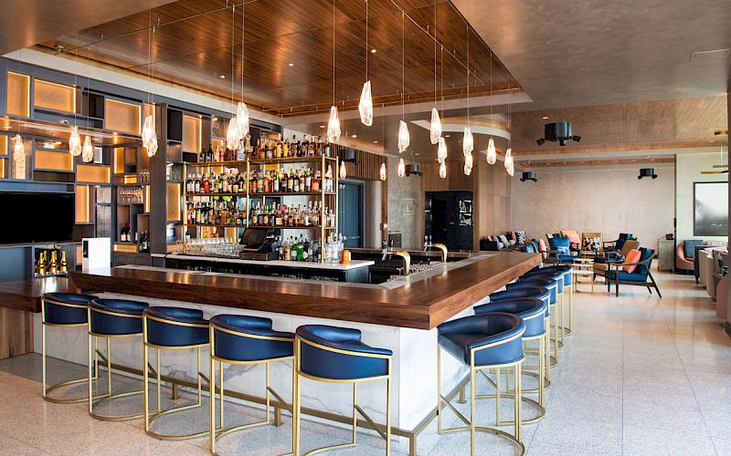 A modern bar with a wooden counter, blue stools, hanging lights, and a well-stocked shelf of bottles. The setting is contemporary and inviting.