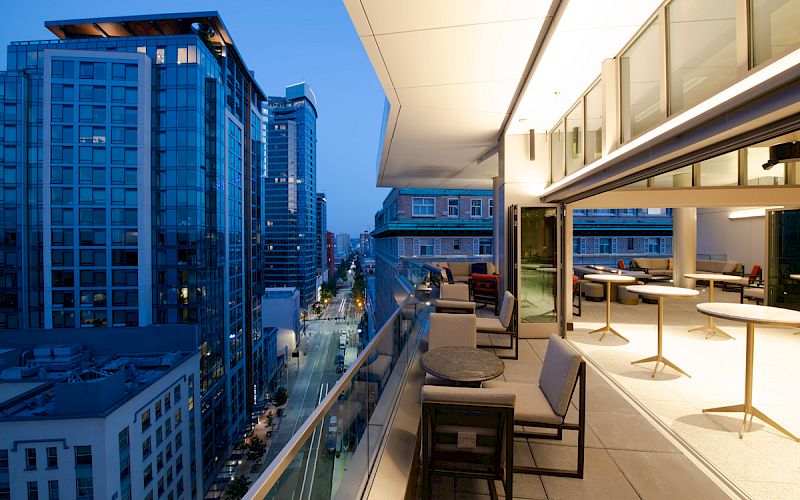The image shows a modern outdoor balcony with seating, tables, and city skyline views. Large windows provide a view into a sleek interior space.