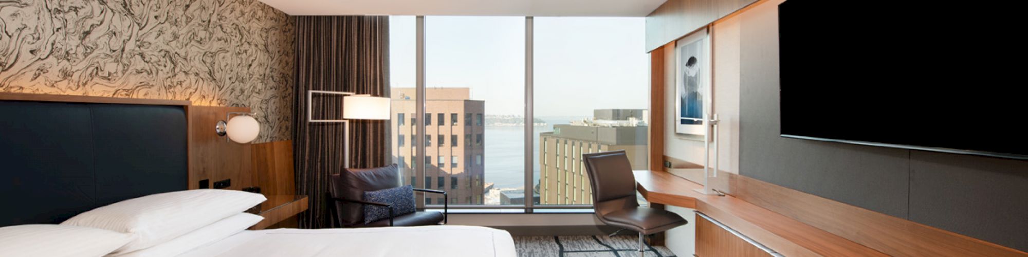This image shows a modern hotel room with a large bed, flat-screen TV, desk, chair, and a window offering a city and waterfront view.