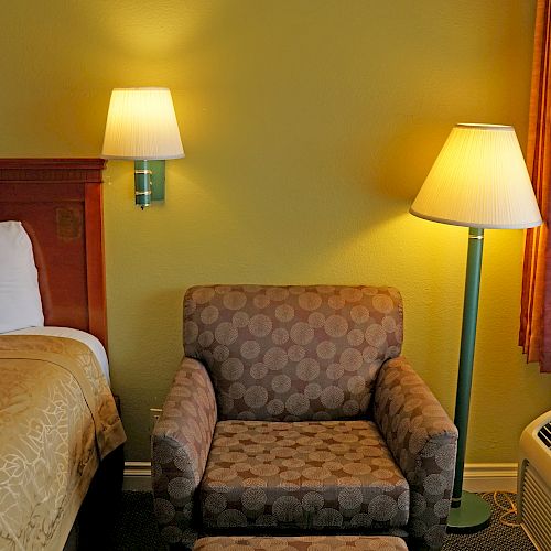 A cozy hotel room featuring a bed, a cushioned chair with an ottoman, two lamps, and a window with curtains nearby.