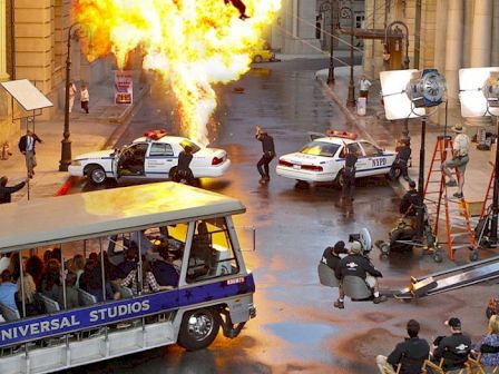 The image depicts a film set with an explosion, police cars, crew, and a Universal Studios tour tram with onlookers.