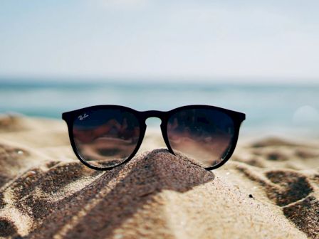 A pair of black sunglasses is placed on sandy ground with a blurred background showing the blue ocean and a clear sky.