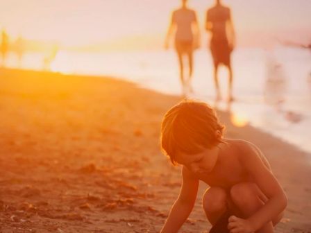 A child plays on the beach at sunset, while two adults walk in the background along the shoreline.