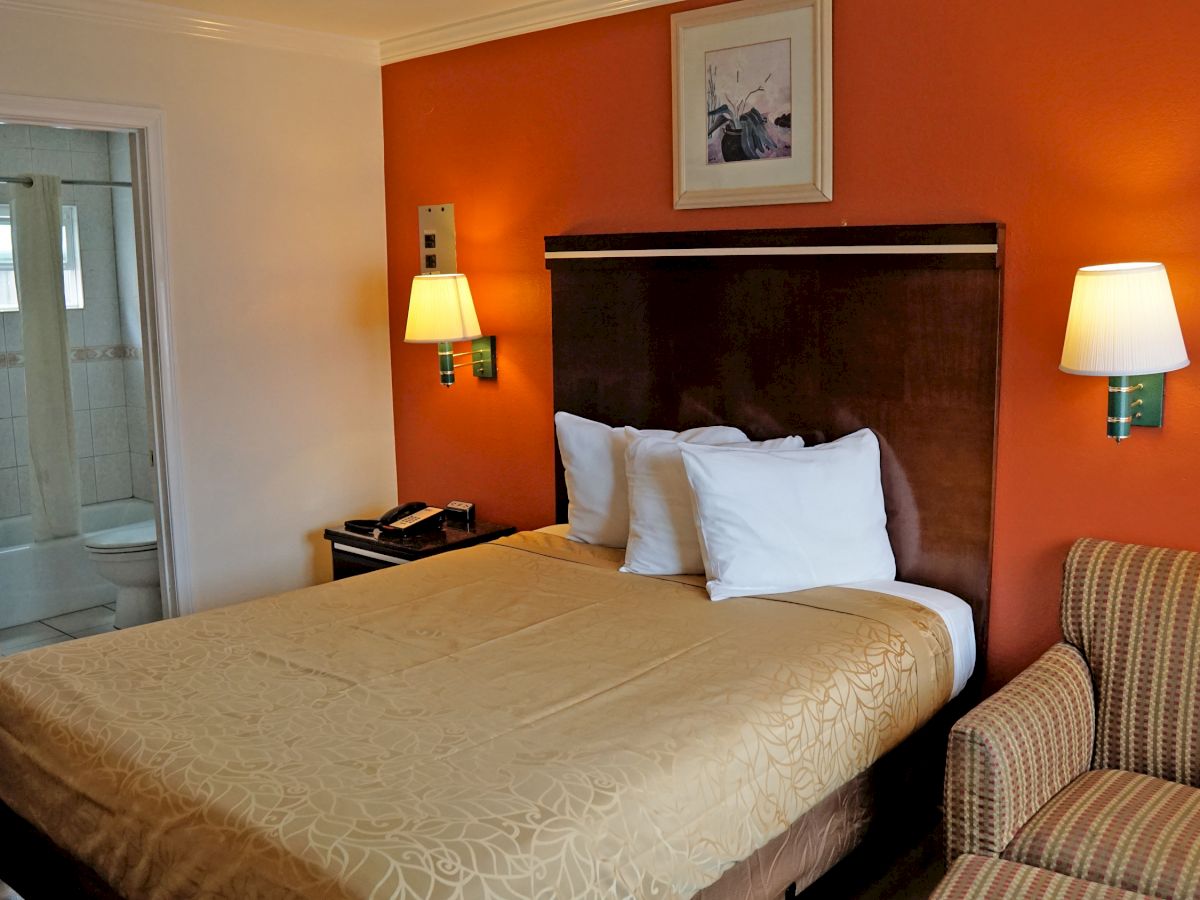 The image shows a cozy hotel room with a bed, nightstands, lamps, a picture on the wall, a chair, and a mirrored closet beside a bathroom door.