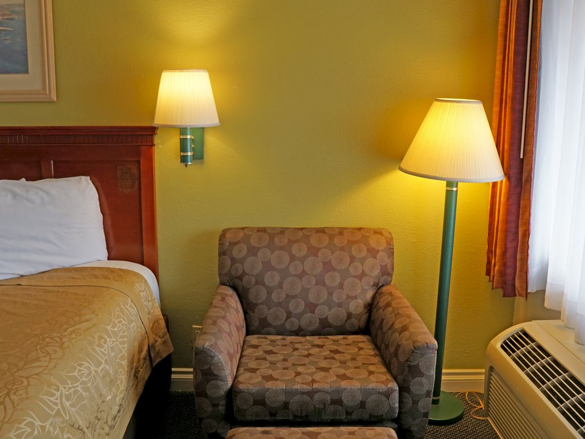 The image shows a cozy corner of a hotel room with a bed, cushioned chair, wall and floor lamps, an air conditioner, and a window with curtains.