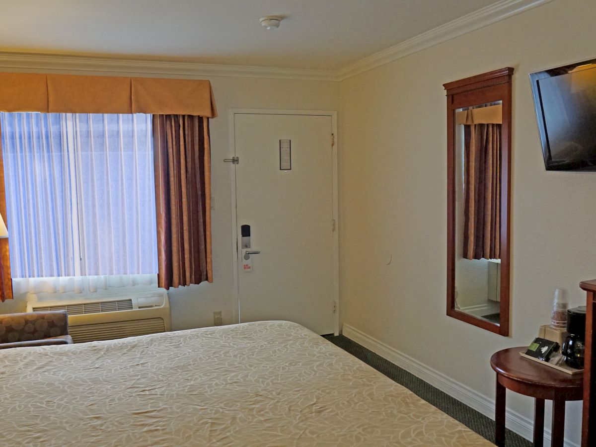 The image shows a tidy hotel room with a large bed, window, wall-mounted TV, mirror, and small desk. The room has warm lighting and simple decor.